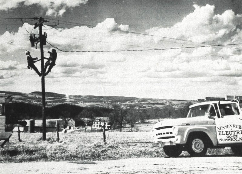 Black-and-white photo of two linemen climbing a pole to reach a pole-mounted transformer. A Sussex Rural Electric Cooperative-branded Ford F100 truck sits at the edge of frame. Source: Sussex Rural Electric Cooperative’s original newsletter “Watts New on the High Point Circuit”
