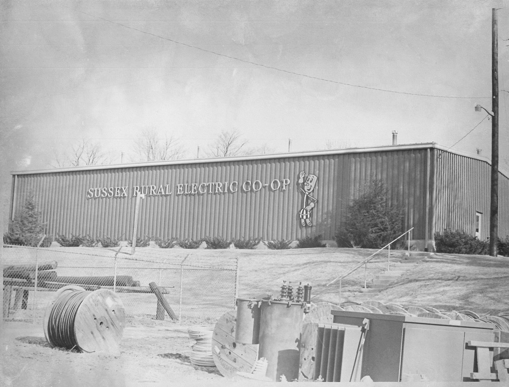 Black-and-white photo of the garage behind Sussex Rural Electric Cooperative's original office, which sports an image of NRECA mascot Willie Wiredhand on its side