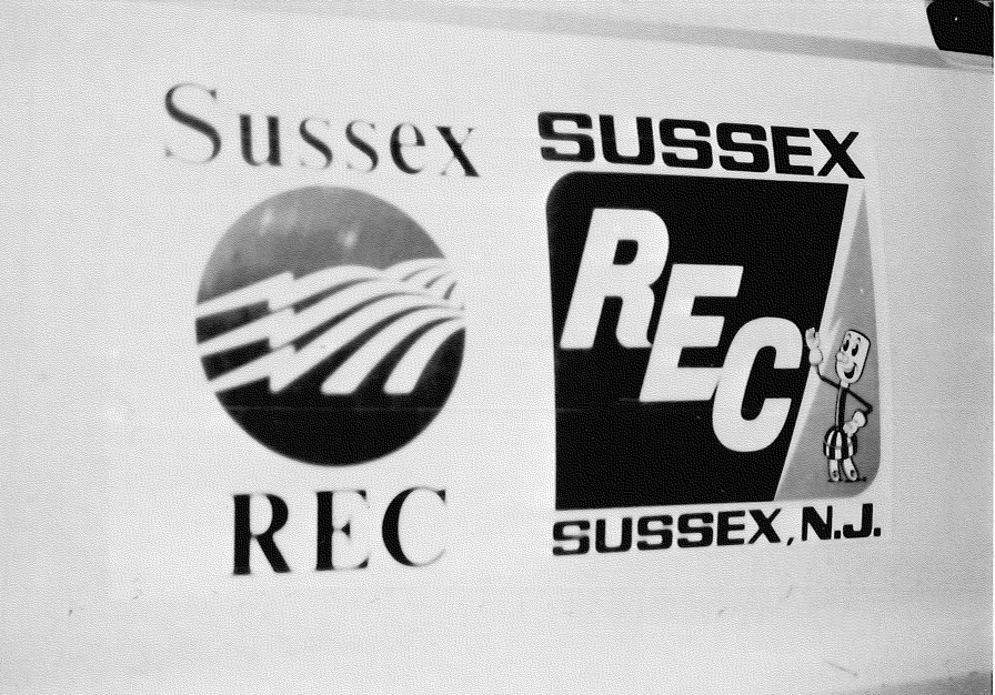A black-and-white, closeup photo of a vintage Sussex Rural Electric Cooperative vehicle door. The door has both the “Sussex REC” found on its vehicles today as well as an image including NRECA mascot Willie Wiredhand.