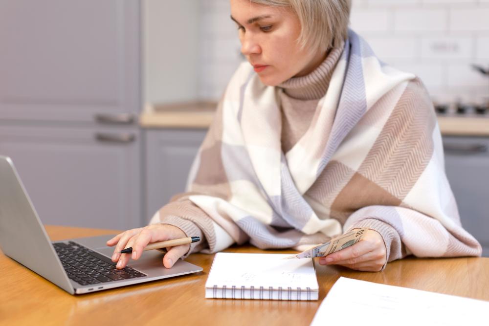 Image of a young woman bundled in a blanket, sitting at a kitchen table with a notepad and a laptop while holding cash