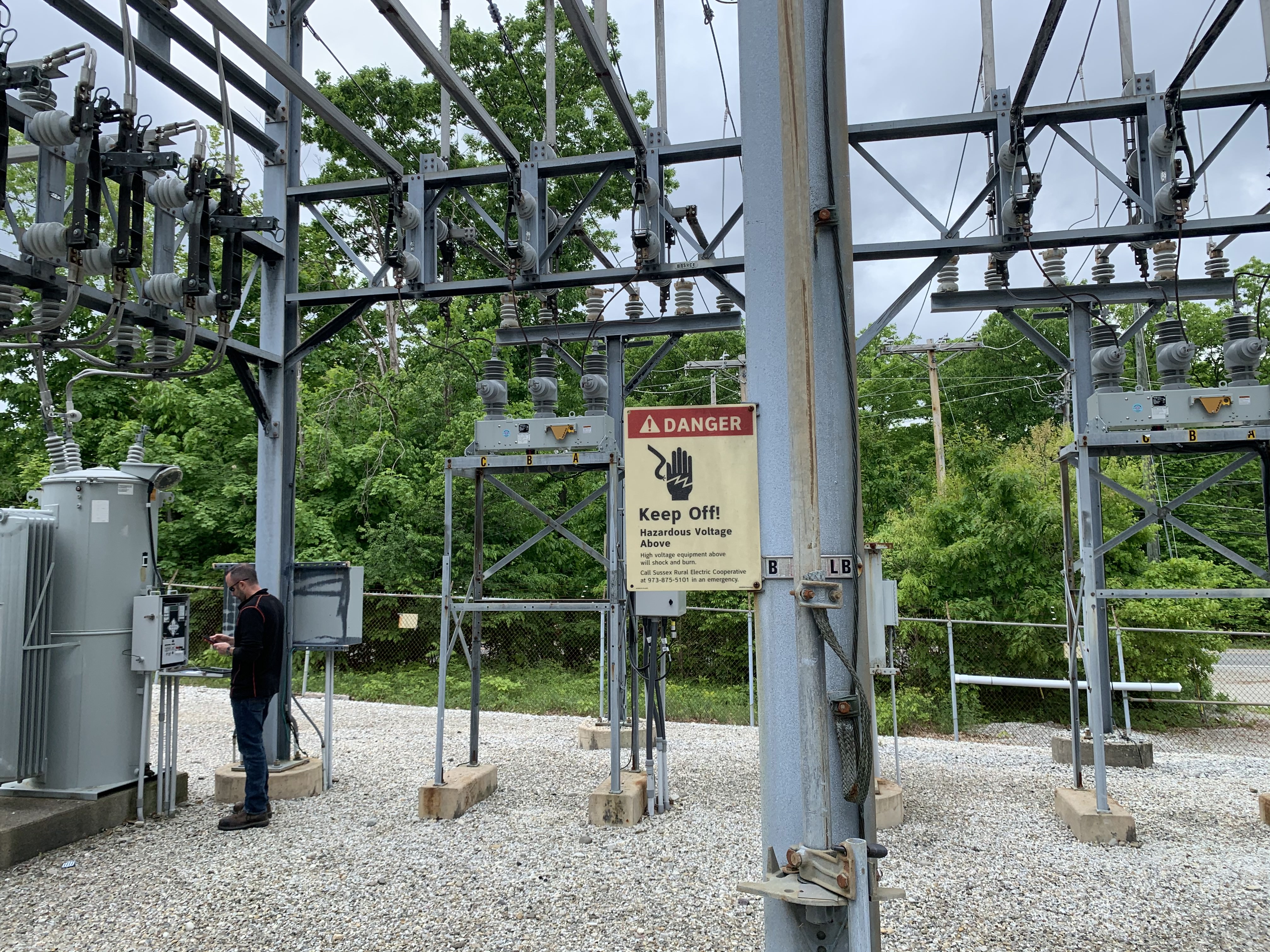 Photo of SREC engineer at substation, with a sign in the foreground that reads: "! DANGER - Keep Off! Hazardous Voltage Above. High voltage and equipment above will shock and burn. Call Sussex Rural Electric Cooperative at 973-875-5101 in an emergency."