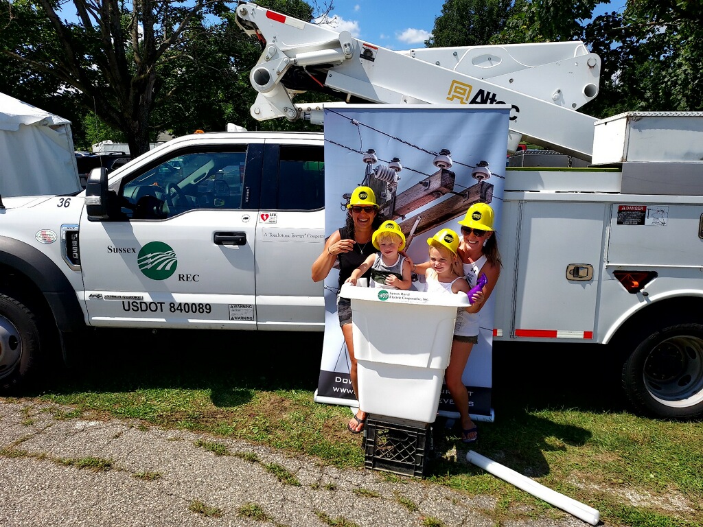 Two women and their children pose with plastic hardhats in a photo booth that includes a mock bucket, a backdrop that shows the top of a utility pole, and a real bucket truck